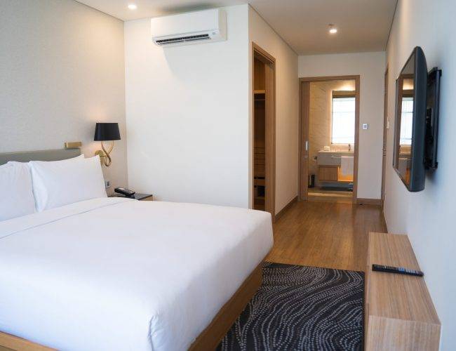 Small hotel room interior with double bed and bathroom. Double bed with white sheets complemented with wall lamp, TV and air conditioner. Hotel room concept